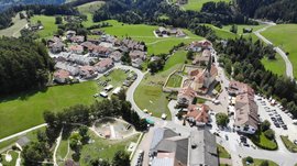 Il Parco Active a Terento L'immagine mostra una foto aerea di un piccolo villaggio situato in un paesaggio collinare verde. Si possono vedere numerose case con tetti rossi, una chiesa con una torre appuntita, nonché un grande parco giochi e un campo sportivo, circondati da prati e boschi.