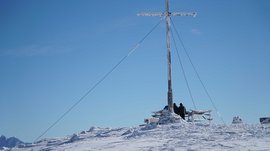 Valles - Jochtal - Steinermandl L'immagine mostra una cima di montagna coperta di neve con una grande croce di vetta parzialmente ricoperta di ghiaccio. Due persone con abbigliamento invernale stanno accanto alla croce e guardano verso l'orizzonte sotto un cielo azzurro e limpido.