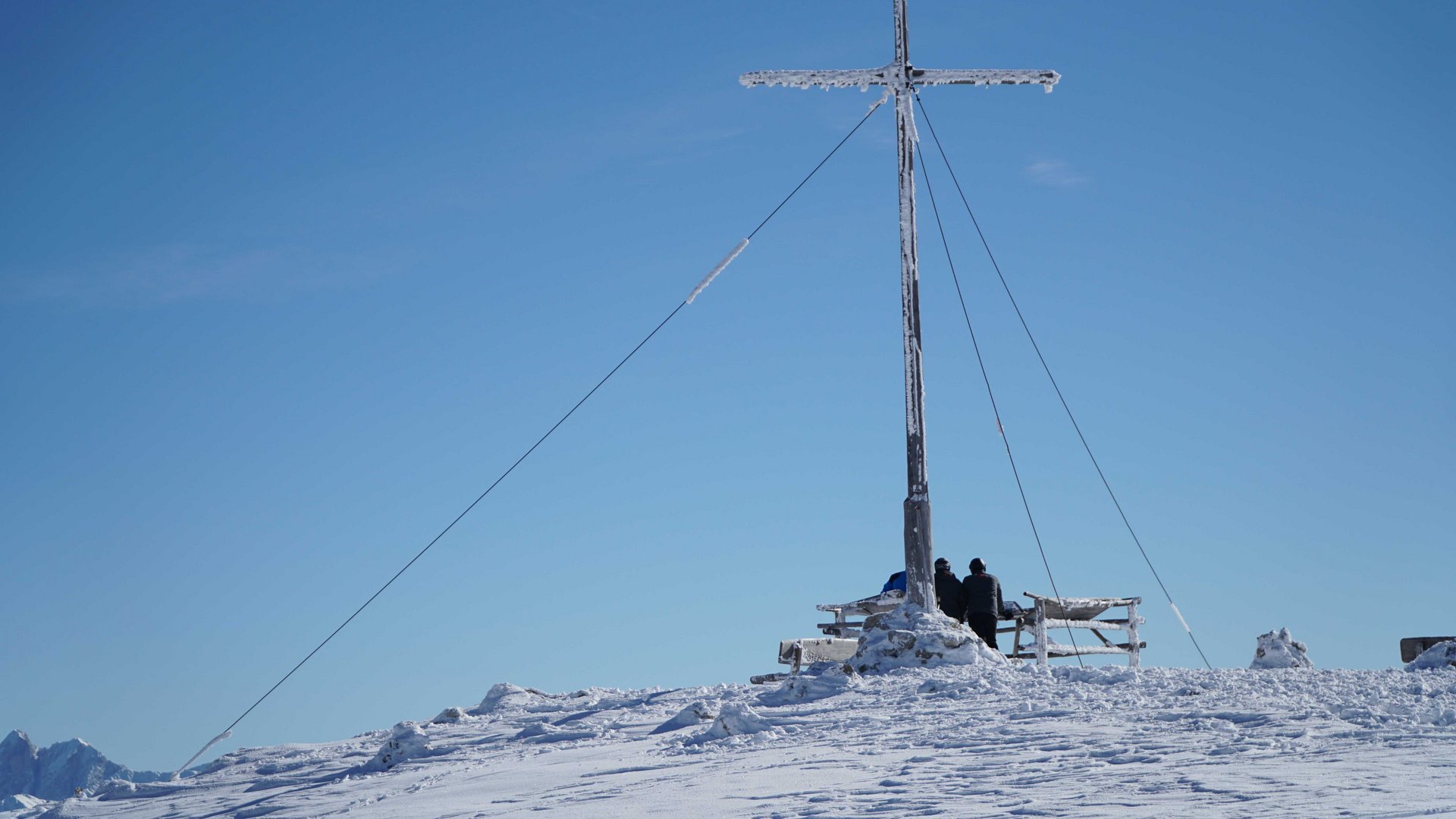 Valles - Jochtal - Steinermandl L'immagine mostra una cima di montagna coperta di neve con una grande croce di vetta parzialmente ricoperta di ghiaccio. Due persone con abbigliamento invernale stanno accanto alla croce e guardano verso l'orizzonte sotto un cielo azzurro e limpido.