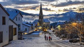L’Avvento di Terento L'immagine mostra una strada di un villaggio innevata con una chiesa sullo sfondo e case decorate in modo festoso. Una famiglia passeggia in un paesaggio invernale pittoresco al tramonto.