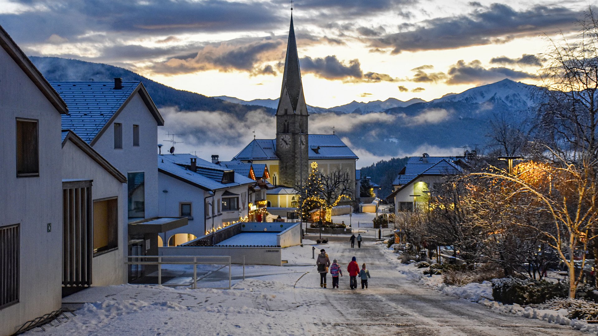 L’Avvento di Terento L'immagine mostra una strada di un villaggio innevata con una chiesa sullo sfondo e case decorate in modo festoso. Una famiglia passeggia in un paesaggio invernale pittoresco al tramonto.