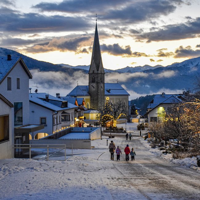 L’Avvento di Terento L'immagine mostra una strada di un villaggio innevata con una chiesa sullo sfondo e case decorate in modo festoso. Una famiglia passeggia in un paesaggio invernale pittoresco al tramonto.