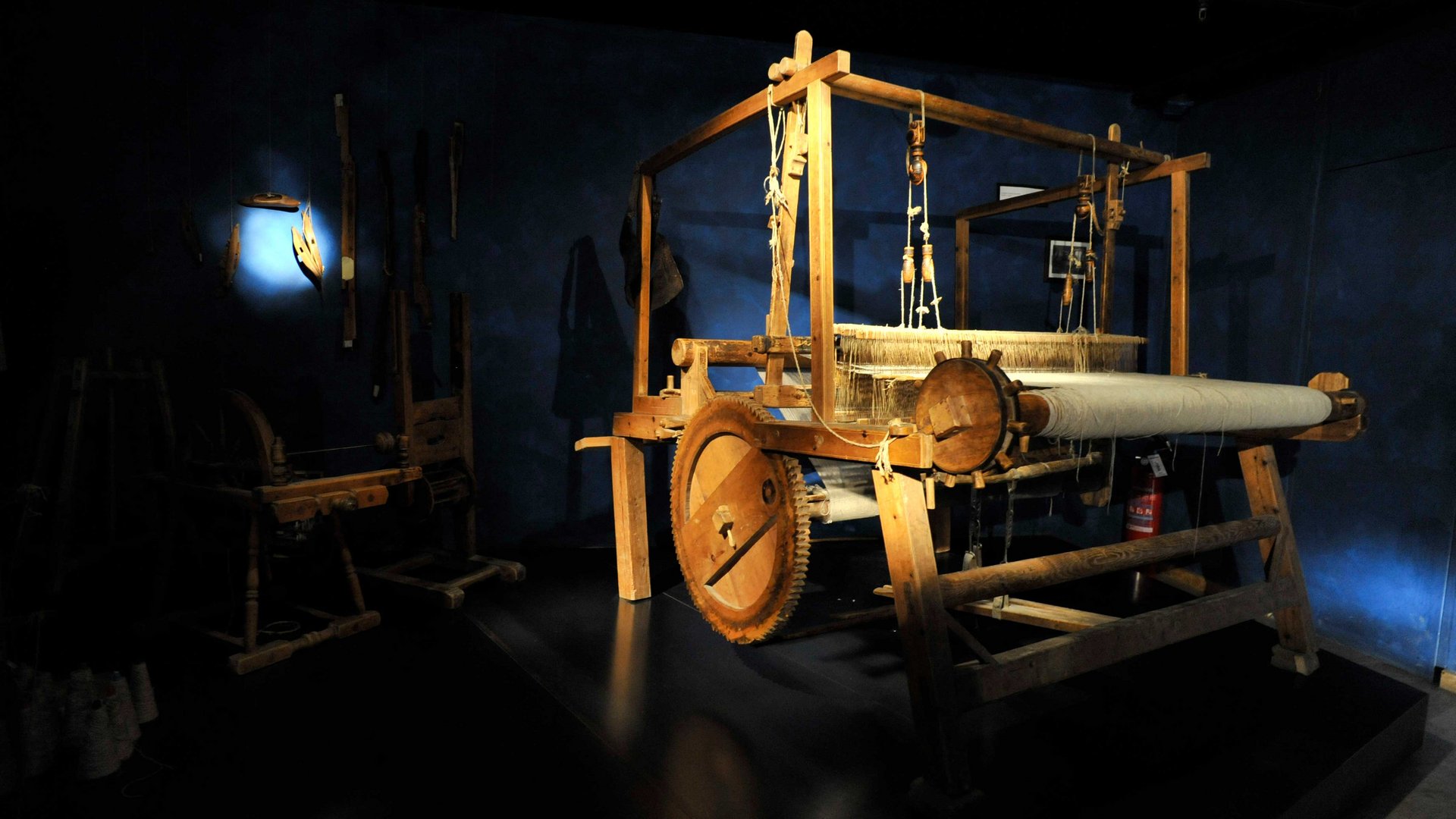 Il Mondo del Loden a Vandoies L'immagine mostra un grande telaio in legno in una stanza buia, illuminata da un raggio di luce. Sullo sfondo si riconoscono altri utensili storici appesi al muro.