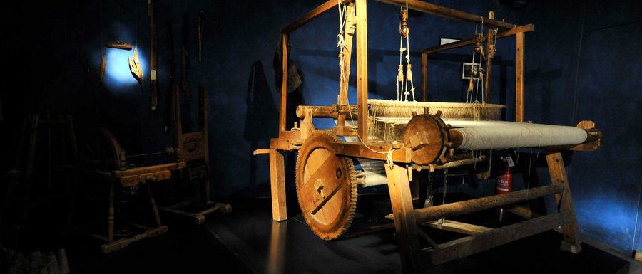 Le località dell’area vacanze Rio Pusteria L'immagine mostra un grande telaio in legno in una stanza buia, illuminata da un raggio di luce. Sullo sfondo si riconoscono altri utensili storici appesi al muro.