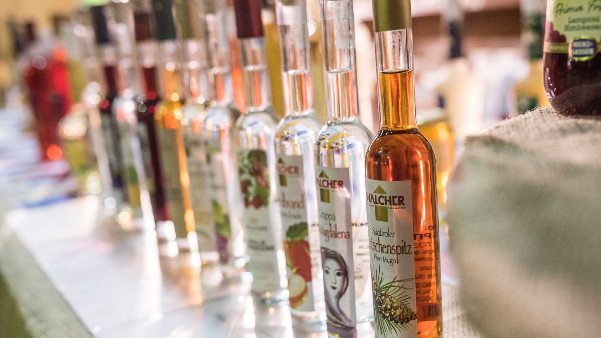 Shopping a Rio di Pusteria (BZ) L'immagine mostra una fila di bottiglie con vari liquori, disposte fianco a fianco. Ogni bottiglia ha un'etichetta con motivi e testi diversi che indicano sapori o ingredienti differenti.