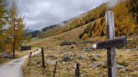 La Valle di Altafossa, presso Maranza L'immagine mostra un sentiero escursionistico che attraversa un paesaggio autunnale con alberi colorati, in parte gialli e arancioni. Il cielo è nuvoloso e sul bordo del sentiero si trova un cartello di legno con la scritta "Großberghütte". Sullo sfondo si vedono alcune baite e una collina coperta di boschi.