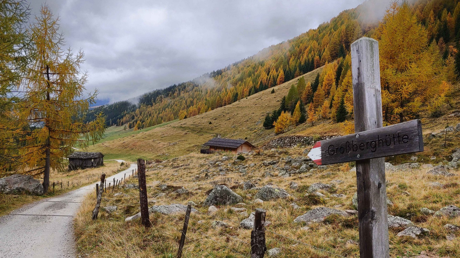 La Valle di Altafossa, presso Maranza L'immagine mostra un sentiero escursionistico che attraversa un paesaggio autunnale con alberi colorati, in parte gialli e arancioni. Il cielo è nuvoloso e sul bordo del sentiero si trova un cartello di legno con la scritta "Großberghütte". Sullo sfondo si vedono alcune baite e una collina coperta di boschi.