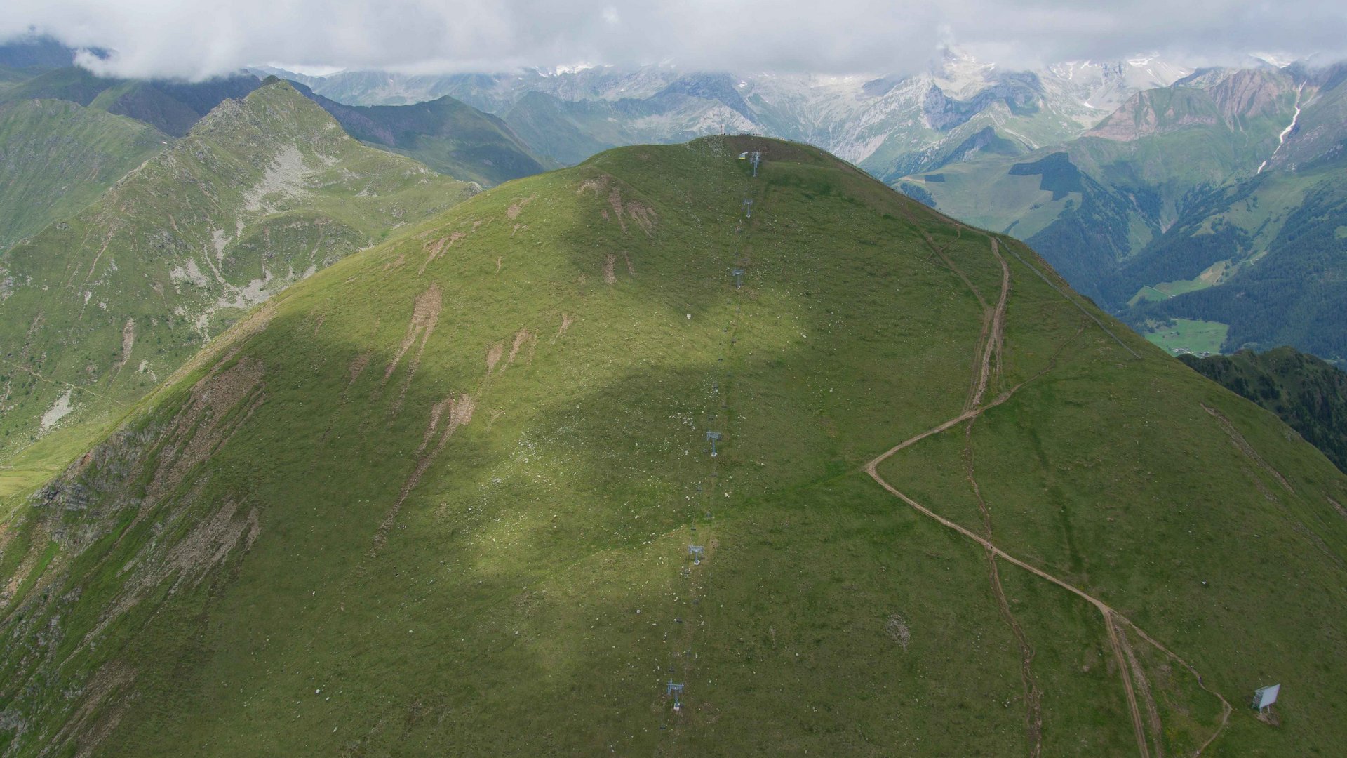 La piattaforma panoramica Gitschberga a Maranza (BZ) L'immagine mostra una cresta montuosa verde coperta d'erba con un sentiero stretto che sale lungo il pendio. Sullo sfondo si vedono altre montagne e vallate, in parte coperte da nuvole.