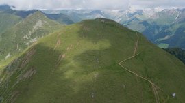 La piattaforma panoramica Gitschberga a Maranza (BZ) L'immagine mostra una cresta montuosa verde coperta d'erba con un sentiero stretto che sale lungo il pendio. Sullo sfondo si vedono altre montagne e vallate, in parte coperte da nuvole.