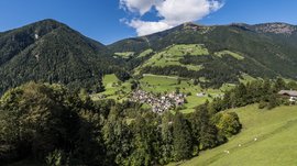 Quando si dice idilliaco… ecco a voi Fundres L'immagine mostra una valle verde e collinare con un piccolo villaggio al centro, circondato da fitte foreste e alte montagne. La scena è tranquilla e idilliaca sotto un cielo azzurro brillante con alcune nuvole bianche.