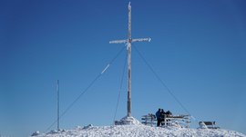 Valles - Jochtal - Steinermandl L'immagine mostra una grande croce di vetta, coperta di neve e ghiaccio, sotto un cielo azzurro brillante. Sullo sfondo ci sono persone accanto a una panchina, che godono della vista invernale dalla cima della montagna.