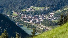 Alla scoperta di Rio di Pusteria L'immagine mostra una piccola città che si estende in una valle verde, circondata da colline densamente boscose. Nel centro della città spicca un campanile, e in primo piano è visibile un lago, sulle cui rive si trova un edificio.