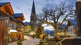 L’Avvento di Terento L'immagine mostra una piazza decorata festosamente con una chiesa sullo sfondo. Un grande albero avvolto da luci e un albero di Natale addobbato si trovano tra le case, anch'esse decorate con catene luminose, mentre la neve ricopre il terreno.
