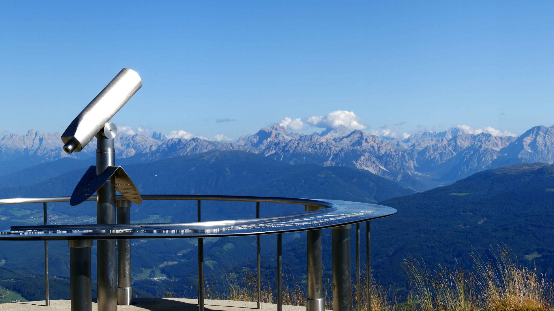 La piattaforma panoramica Gitschberga a Maranza (BZ) L'immagine mostra una piattaforma panoramica per l'osservazione su una cima di montagna, dotata di un cannocchiale fisso. Sullo sfondo si estende una vasta vallata, circondata da numerose catene montuose e cime, sotto un cielo limpido e azzurro.