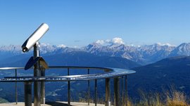 La piattaforma panoramica Gitschberga a Maranza (BZ) L'immagine mostra una piattaforma panoramica per l'osservazione su una cima di montagna, dotata di un cannocchiale fisso. Sullo sfondo si estende una vasta vallata, circondata da numerose catene montuose e cime, sotto un cielo limpido e azzurro.