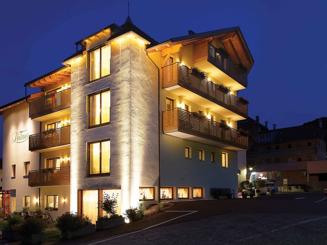 Benvenuti nell’Area Vacanze Sci e Malghe Rio Pusteria! L'immagine mostra un edificio alberghiero illuminato e su più piani durante la notte. La facciata dell'edificio è illuminata con una luce giallastra e sui balconi ci sono fioriere. L'edificio emana un'atmosfera calda e accogliente, mentre sullo sfondo si vede un cielo notturno scuro.