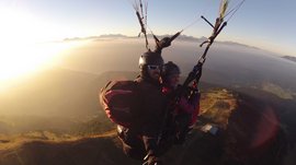 Parapendio in Alto Adige L'immagine mostra due persone che fluttuano nell'aria durante il parapendio, mentre il sole è basso nel cielo e immerge il paesaggio in una luce dorata. Sullo sfondo si vedono montagne e un'ampia pianura che svanisce in una dolce nebbia.