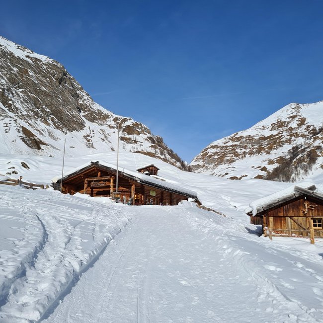 Kuttnhütte L'immagine mostra un rifugio di montagna in legno, circondato da neve profonda, su un pendio innevato. Sullo sfondo si ergono montagne rocciose coperte di neve sotto un cielo limpido e blu.