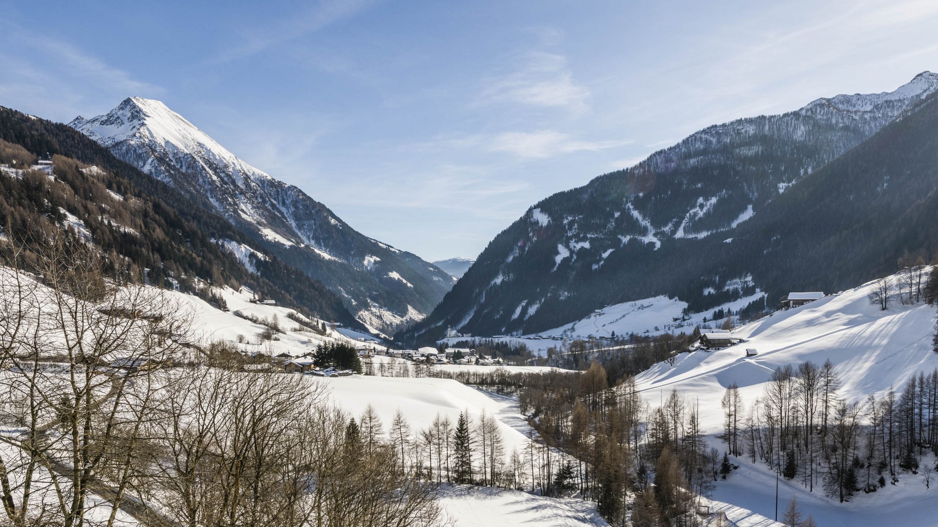 Quando si dice idilliaco… ecco a voi Fundres L'immagine mostra un paesaggio montano invernale con pendii coperti di neve e una valle al centro. Il sole splende, il cielo è limpido e azzurro, e in primo piano si vedono alberi spogli. Le montagne sullo sfondo sono anch'esse coperte di neve, il che trasmette un'atmosfera di pace e tranquillità.