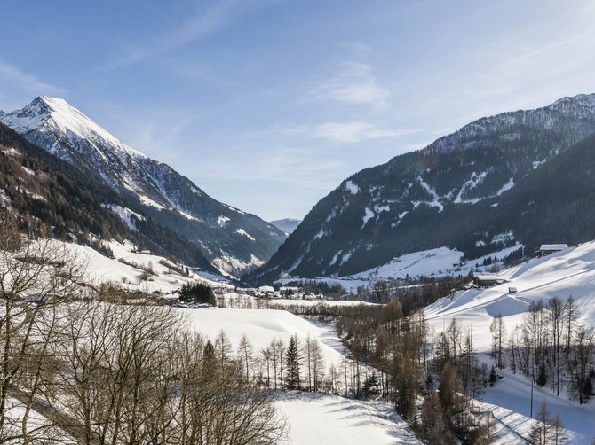 Le località dell’area vacanze Rio Pusteria L'immagine mostra un paesaggio montano invernale con pendii coperti di neve e una valle al centro. Il sole splende, il cielo è limpido e azzurro, e in primo piano si vedono alberi spogli. Le montagne sullo sfondo sono anch'esse coperte di neve, il che trasmette un'atmosfera di pace e tranquillità.