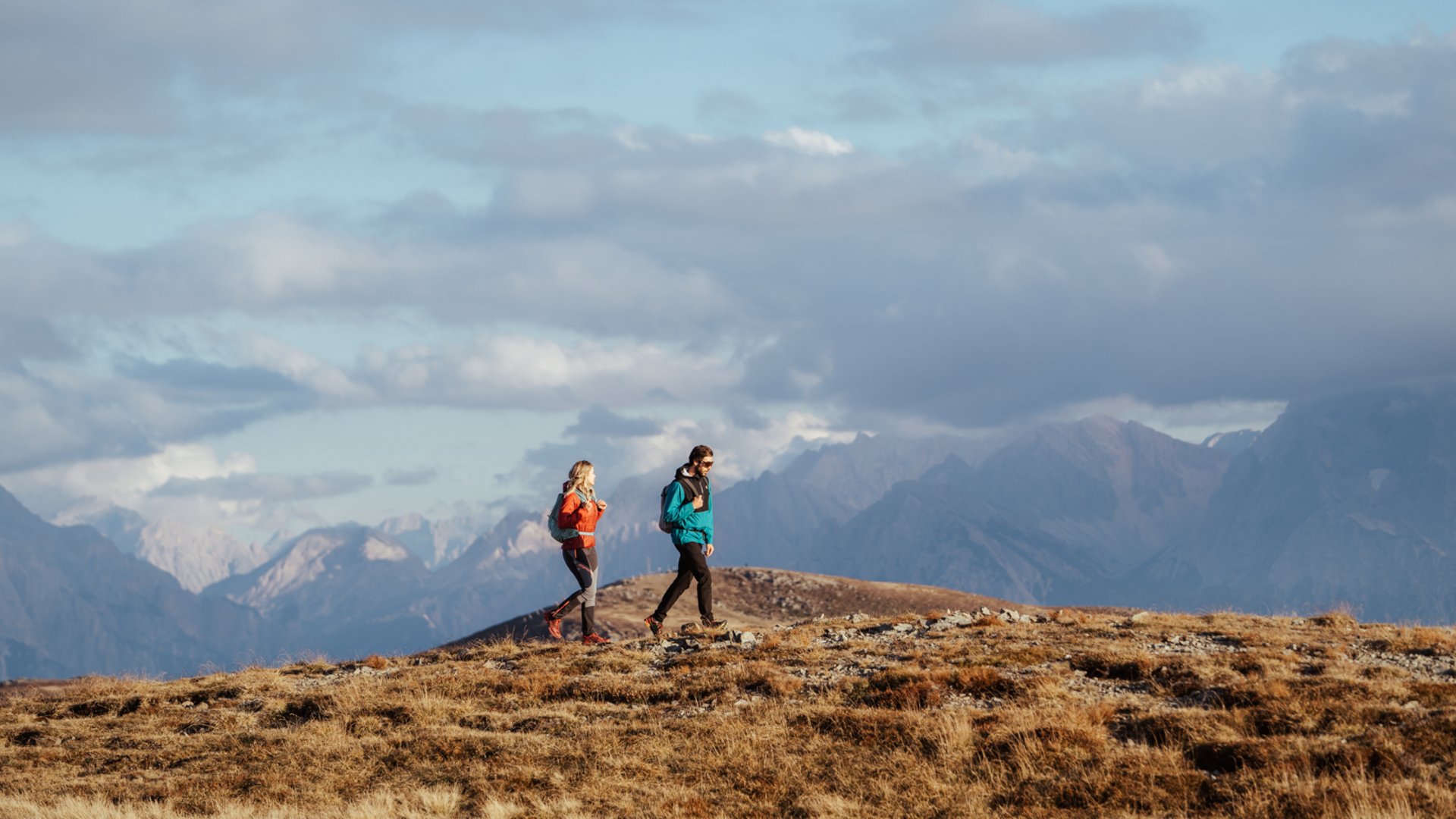 I percorsi più belli delle Dolomiti L'immagine mostra due escursionisti, un uomo e una donna, che camminano con abiti caldi e zaini attraverso un paesaggio montano erboso. Sullo sfondo si vedono alte montagne, in parte coperte da nuvole.