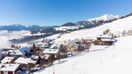 Vacanze sugli sci con i bambini a Terento L'immagine mostra un villaggio innevato tra le montagne, con tetti e colline coperti di neve. Sullo sfondo si vedono colline boschive e maestose montagne sotto un cielo azzurro e limpido.