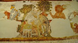 Alla scoperta di Castel Rodengo L'immagine mostra un antico affresco murale parzialmente danneggiato. Raffigura una scena con un cavaliere in armatura, un albero e una figura mitologica con capelli rossi. I colori sono sbiaditi e alcune parti dell'affresco mancano.