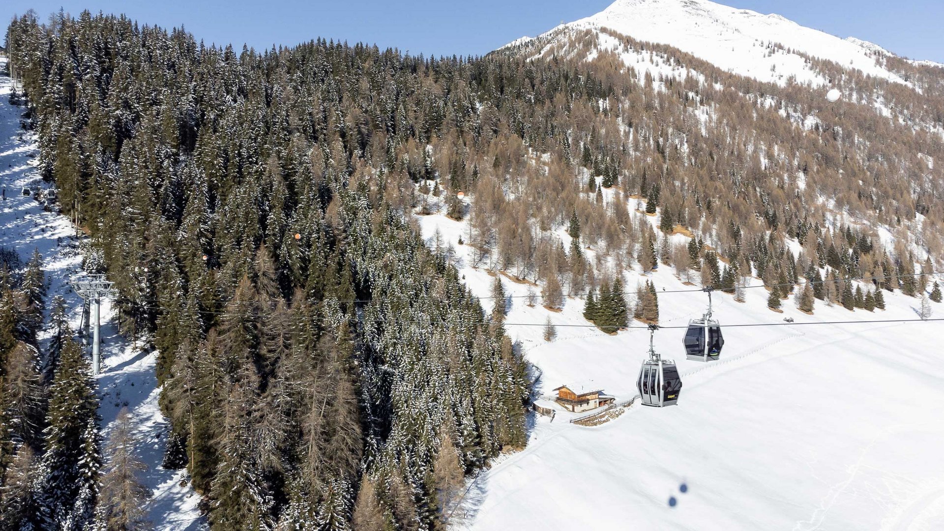 La Valle di Altafossa, presso Maranza L'immagine mostra una funivia con cabine che viaggia sopra un paesaggio innevato. Sullo sfondo si vedono foreste fitte coperte di neve e una cima di montagna alta e innevata. Alla base del pendio c'è una piccola baita. Il cielo è limpido e azzurro.