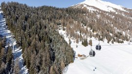 La Valle di Altafossa, presso Maranza L'immagine mostra una funivia con cabine che viaggia sopra un paesaggio innevato. Sullo sfondo si vedono foreste fitte coperte di neve e una cima di montagna alta e innevata. Alla base del pendio c'è una piccola baita. Il cielo è limpido e azzurro.