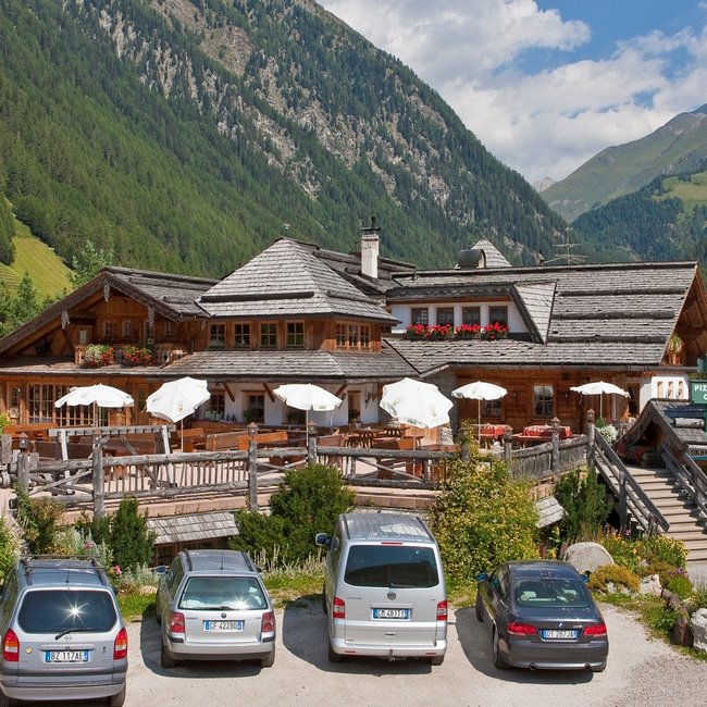 Restaurant Pizzeria Die Mühle L'immagine mostra un grande hotel di montagna rustico con tetto in legno e pietra, circondato da montagne verdi e foreste. Davanti all'hotel c'è una terrazza con ombrelloni bianchi e diverse auto parcheggiate, e un cartello indica una pizzeria, un caffè e una scuola di sci.