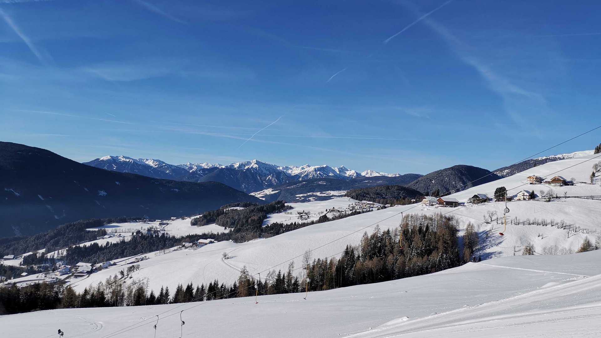 Vacanze sugli sci con i bambini a Terento L'immagine mostra un paesaggio montano coperto di neve con un cielo azzurro limpido. In primo piano si vedono impianti di risalita e pendii, mentre sullo sfondo si estendono diverse catene montuose.