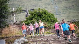 Il Parco del Sole Gitschberg – Maranza L'immagine mostra un gruppo di bambini che stanno salendo un sentiero roccioso. Sullo sfondo si vede una grande scultura di un'aquila con le ali spiegate, nascosta dietro un cespuglio.