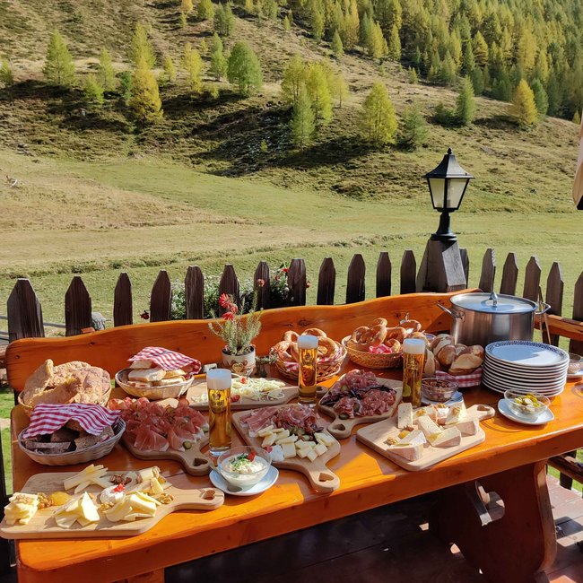 Pranter Stadelhütte L'immagine mostra un tavolo rustico di legno all'aperto, apparecchiato con una varietà di cibi, tra cui formaggio, pane, affettati e pretzel. In lontananza si vede un paesaggio pittoresco con colline verdi e alcuni alberi sparsi.