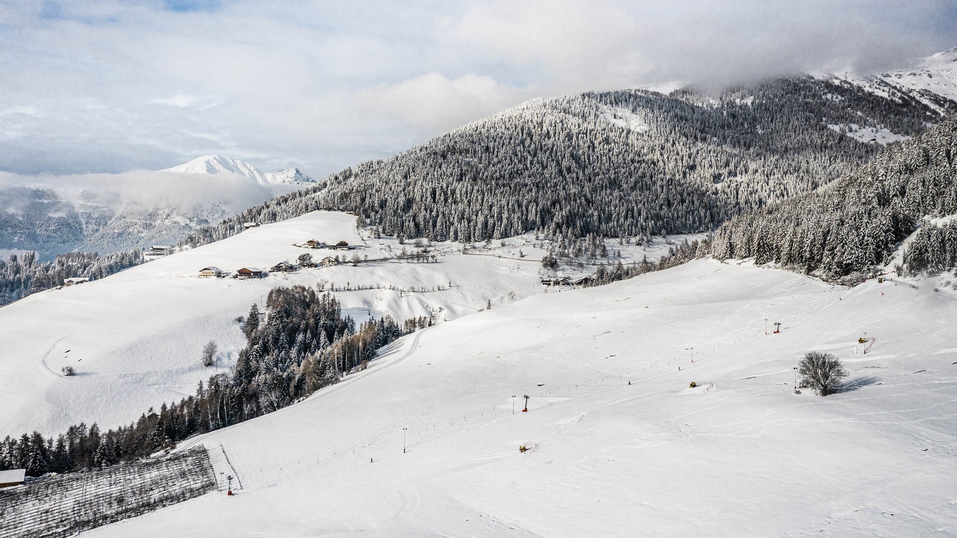 Vacanze sugli sci con i bambini a Terento L'immagine mostra un paesaggio ampio e coperto di neve con dolci colline e una fitta foresta sullo sfondo. Sulle colline si vedono case sparse e impianti di risalita per sci, mentre le montagne sullo sfondo sono parzialmente coperte da nuvole.