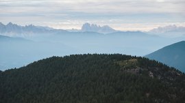 Valles - Jochtal - Steinermandl L'immagine mostra un paesaggio collinare con montagne densamente boschive in primo piano. Sullo sfondo sono visibili diverse catene montuose che appaiono in varie tonalità di blu, poiché sono avvolte dalla nebbia o foschia, mentre il cielo è nuvoloso.