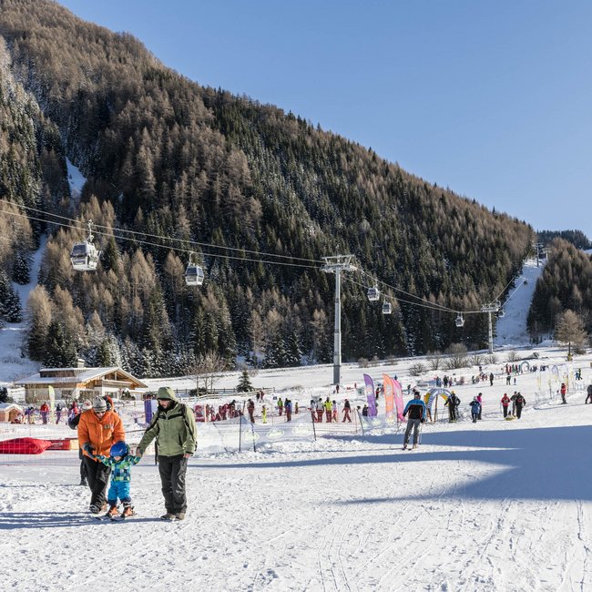 Benvenuti nell’Area Vacanze Sci e Malghe Rio Pusteria! Stazione sciistica con persone, seggiovie e montagne innevate sotto cielo sereno