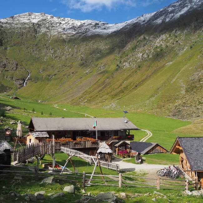 Wieserhütte L'immagine mostra una baita in un verde prato di una valle circondata da montagne. Davanti alla baita ci sono recinzioni di legno, un piccolo ponte e diverse biciclette. Le montagne circostanti hanno alcune cime innevate, e sopra si estende un cielo azzurro e limpido.