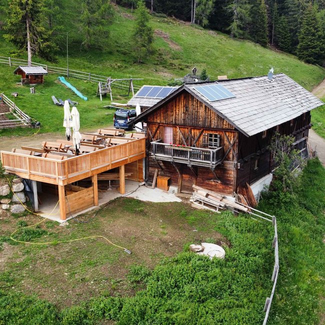 Pertinger Alm L'immagine mostra un rustico chalet di montagna in legno con una grande terrazza e balconi, circondato da boschi e prati. Davanti allo chalet ci sono tavoli e sedie, e sul tetto sono visibili pannelli solari.