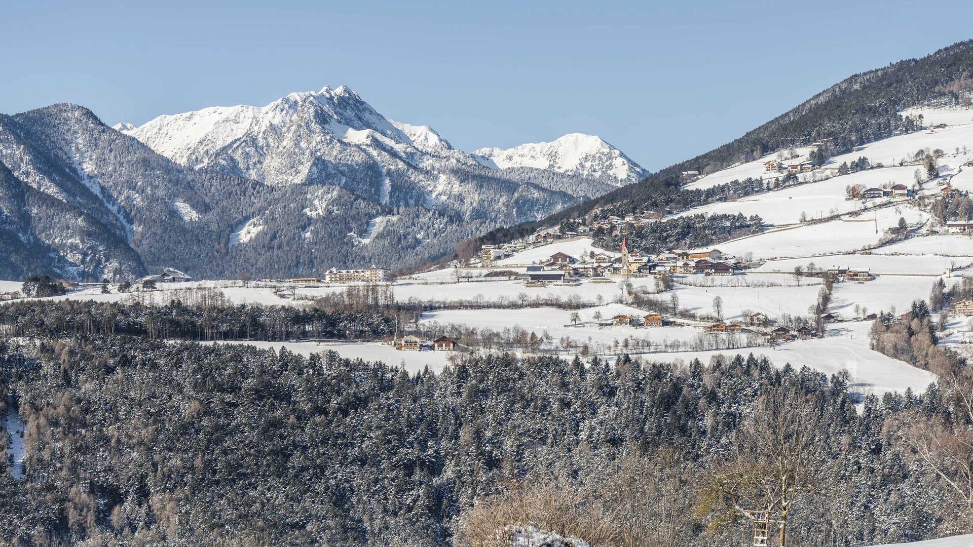 La frazione montana di Spinga L'immagine mostra un paesaggio innevato con un piccolo villaggio in primo piano, circondato da campi e foreste innevate. Sullo sfondo si ergono montagne impressionanti con cime innevate sotto un cielo blu limpido.
