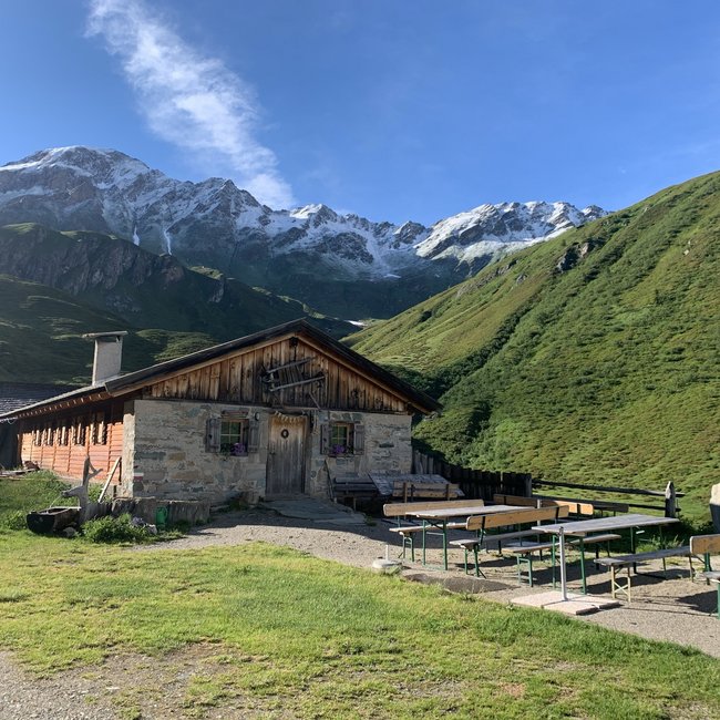 Weitenbergalm L'immagine mostra un rustico rifugio di montagna in pietra e legno in una valle verde, circondato da alte montagne con cime innevate. Davanti al rifugio ci sono diverse panche e tavoli per la birra vuoti, e il cielo è limpido e blu.