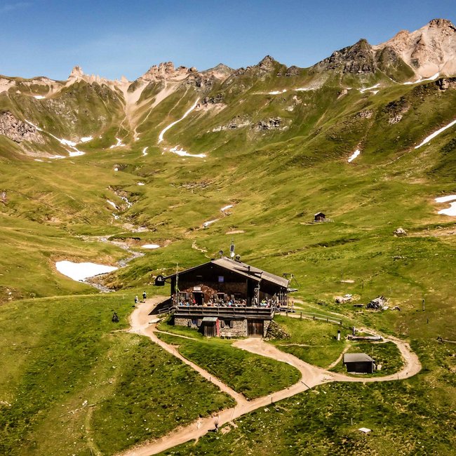 Brixner Hütte L'immagine mostra un baita alpino in mezzo a un paesaggio montano verde con alcune macchie di neve sparse. Sullo sfondo si ergono montagne ripide e rocciose sotto un cielo azzurro e limpido. Un piccolo ruscello scorre attraverso la valle accanto al baita.