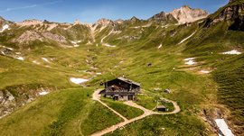 L’Alta via di Fundres L'immagine mostra un baita alpino in mezzo a un paesaggio montano verde con alcune macchie di neve sparse. Sullo sfondo si ergono montagne ripide e rocciose sotto un cielo azzurro e limpido. Un piccolo ruscello scorre attraverso la valle accanto al baita.