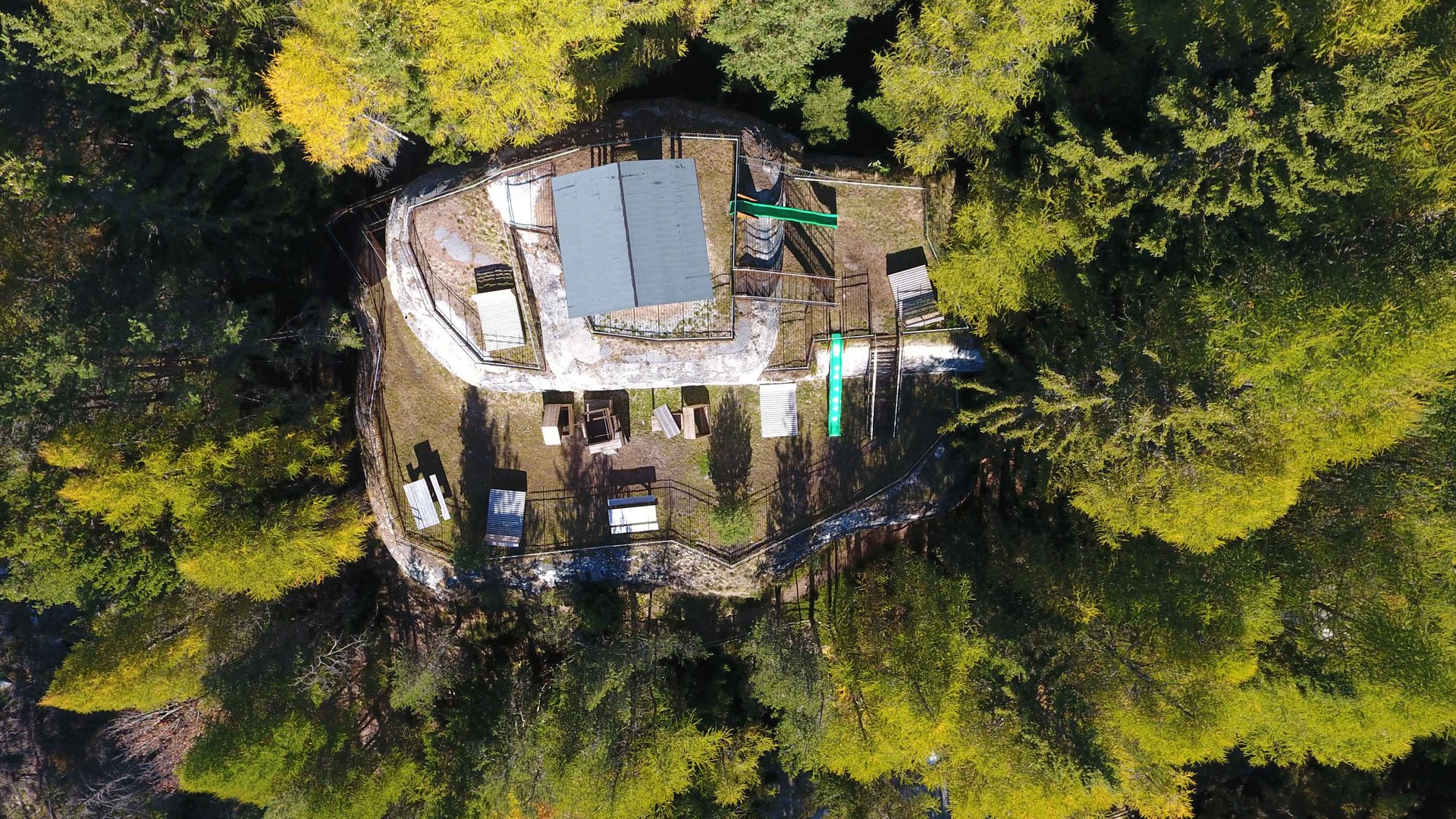 La zona ricreativa bunker di Spinga L'immagine mostra una foto aerea di un edificio situato nel mezzo di una fitta foresta. Intorno all'edificio sono disposte diverse panchine e tavoli, così come attrezzature da gioco, e l'ambiente appare tranquillo e naturale.