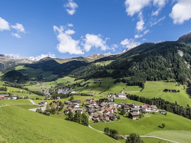 Le località dell’area vacanze Rio Pusteria L'immagine mostra un paesaggio alpino pittoresco con un piccolo villaggio circondato da prati verdi e colline boschive. Sullo sfondo si vedono montagne innevate sotto un cielo blu limpido con alcune nuvole.