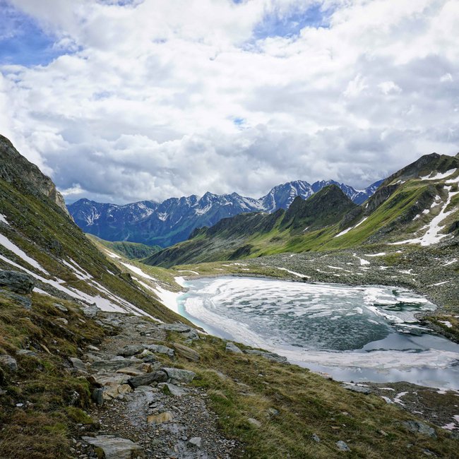 Trovate il vostro hotel in Val Pusteria L'immagine mostra un paesaggio montano con cime innevate sullo sfondo. Un lago parzialmente ghiacciato si trova in una valle, circondato da sentieri rocciosi e pendii coperti di erba.