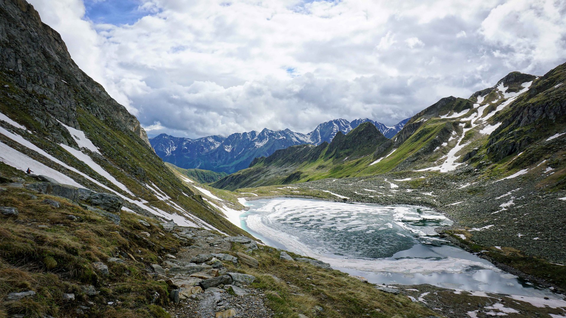 L’Alta via di Fundres L'immagine mostra un paesaggio montano con cime innevate sullo sfondo. Un lago parzialmente ghiacciato si trova in una valle, circondato da sentieri rocciosi e pendii coperti di erba.