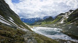 L’Alta via di Fundres L'immagine mostra un paesaggio montano con cime innevate sullo sfondo. Un lago parzialmente ghiacciato si trova in una valle, circondato da sentieri rocciosi e pendii coperti di erba.