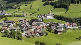 Quando si dice idilliaco… ecco a voi Fundres L'immagine mostra un piccolo villaggio in un paesaggio verde e collinare. Al centro del villaggio si trova una chiesa con una torre a cipolla rossa, circondata da case tradizionali, e sullo sfondo si estendono prati rigogliosi e foreste dense.