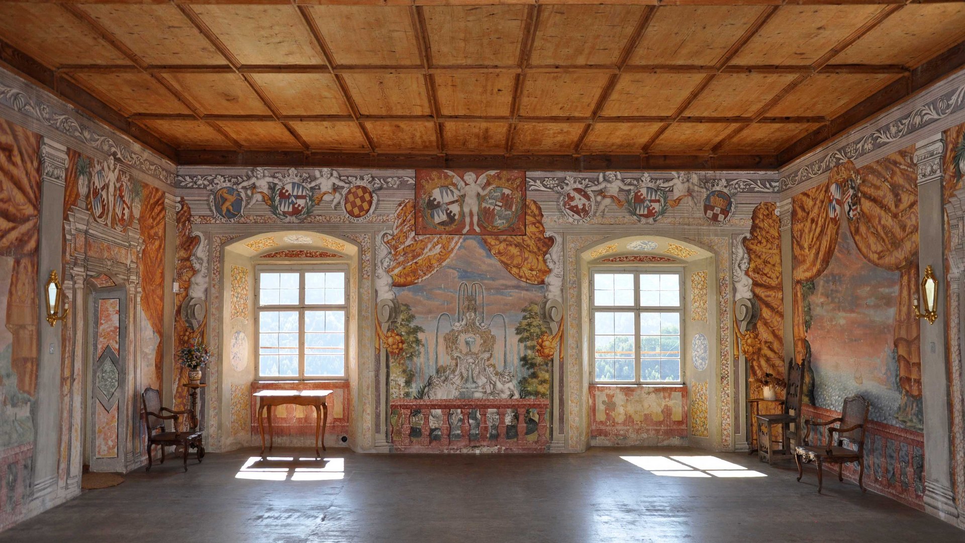 Alla scoperta di Castel Rodengo L'immagine mostra una stanza magnifica con pareti finemente dipinte, che raffigurano scene con angeli, stemmi e paesaggi. La stanza ha un soffitto con travi in legno, due grandi finestre che lasciano entrare la luce ed è arredata con mobili antichi.