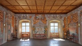 Alla scoperta di Castel Rodengo L'immagine mostra una stanza magnifica con pareti finemente dipinte, che raffigurano scene con angeli, stemmi e paesaggi. La stanza ha un soffitto con travi in legno, due grandi finestre che lasciano entrare la luce ed è arredata con mobili antichi.